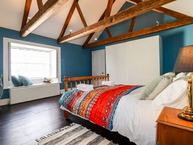 A bedroom with a wooden bed frame colorful bedding a window seat with pillows wooden beams on the ceiling and a white wardrobe at Carew House Cottage in St Day