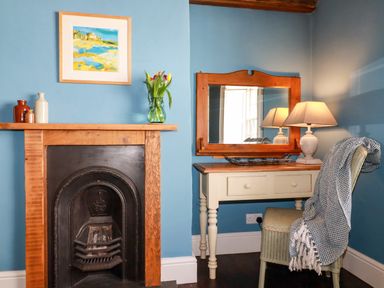 A corner with a fireplace with vases and flowers a dressing table with a mirror lamp and a chair with a blanket at Carew House Cottage in St Day