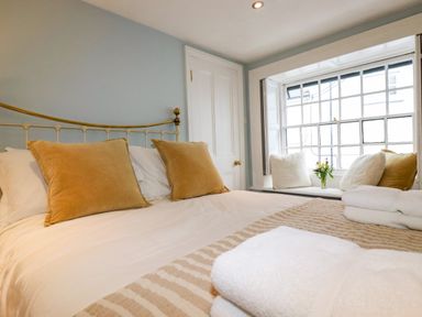 A bedroom with a made bed with white linens and mustard pillows a window seat with cushions and a vase of flowers at Carew House Cottage in St Day