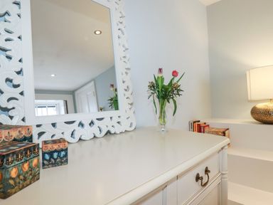 A white dresser with a carved frame mirror a vase with flowers decorative boxes books and a lamp at Carew House Cottage in St Day