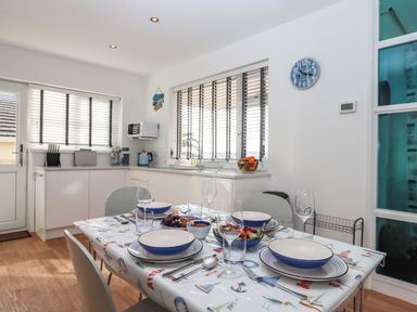 A dining table set with bowls plates glasses and cutlery in a kitchen with white cabinets and windows at Sandy Toes Paignton