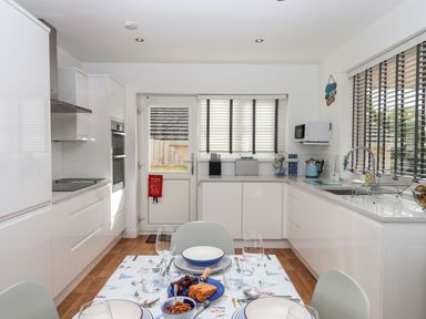 A kitchen with white cabinets a dining table set with plates and glasses a microwave on the wall and windows with blinds at Sandy Toes Paignton