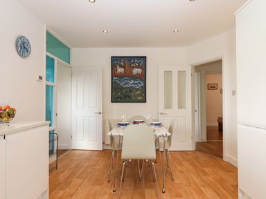 A dining area with a table and four chairs set with dishes and glasses a painting on the wall and white doors at Sandy Toes in Paignton