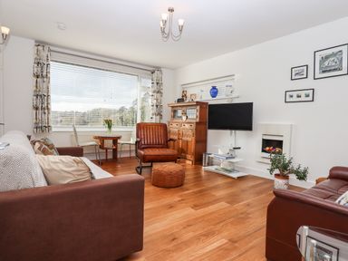 A living room with brown sofas a leather chair a wooden cabinet a television and a small dining table at Sandy Toes in Paignton