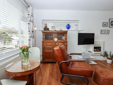 A living room with a leather chair wooden cabinet wall mounted tv and a glass vase with tulips on a table at Sandy Toes in Paignton
