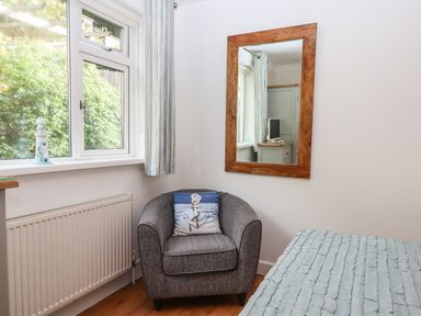 A corner of a bedroom with a grey armchair with an anchor pillow a window a wall mirror and part of a bed at Sandy Toes in Paignton