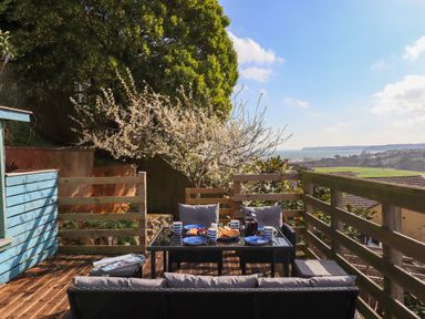 An outdoor wooden deck with a table set for breakfast and cushioned chairs overlooking a coastal view at Sandy Toes in Paignton