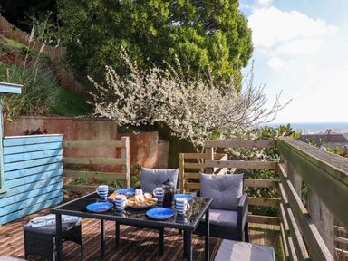 An outdoor deck with a glass table set for tea and cushioned chairs overlooking a garden and distant sea at Sandy Toes in Paignton