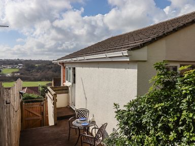 An outdoor wooden deck with a small table and two chairs beside a beige house with a view of fields and houses at Sandy Toes in Paignton