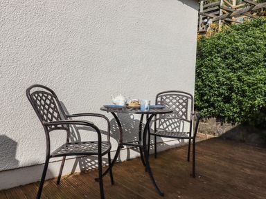 Two metal chairs and a round metal table with tea set on a wooden deck outside next to a white wall and green bushes at Sandy Toes in Paignton