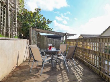 An outdoor wooden deck with a round glass table and four chairs with a fence and plants at Sandy Toes in Paignton