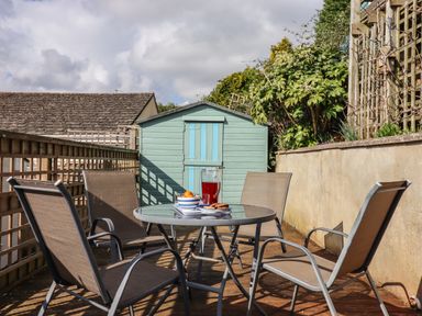 An outdoor patio with a round glass table set with a jug of red drink and snacks surrounded by four chairs and a green shed in the background at Sandy Toes in Paignton