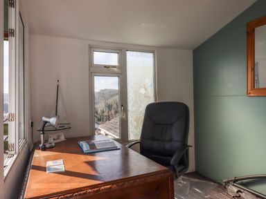 A room with a wooden desk with a lamp, a model sailboat, an open book, and a black office chair near windows at Sandy Toes in Paignton
