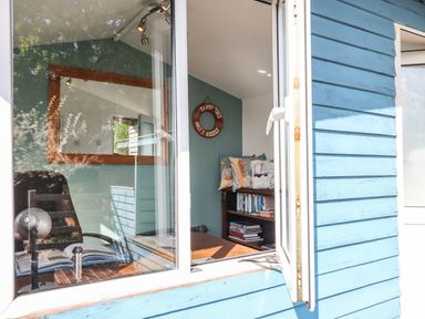 A small room with blue walls featuring a desk with a lamp and open book a bookshelf with books and cushions and a decorative life ring on the wall at Sandy Toes Paignton