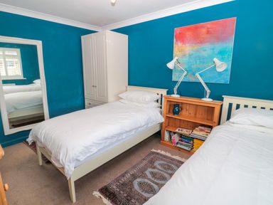 A bedroom with two single beds a wooden bookshelf with lamps and books a white wardrobe and a large mirror on blue walls at Turnstone in Beadnell