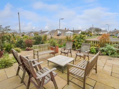 An outdoor patio with wooden chairs and benches arranged around a square table surrounded by plants and houses at Turnstone in Beadnell