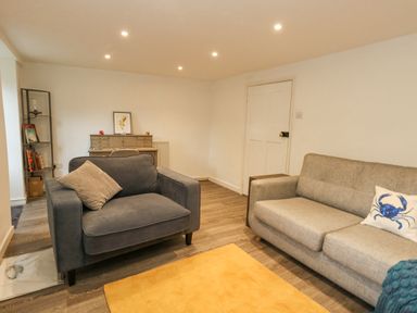 A living room with a gray armchair, gray sofa with a crab pillow, wooden floor, yellow rug, shelving unit, and a desk at Half Pint Cottage in Beaminster