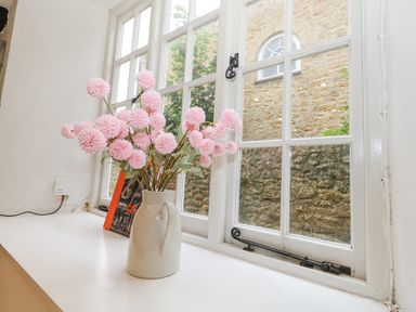 A windowsill with a vase of pink flowers and a book next to a white window with black latches at Half Pint Cottage in Beaminster