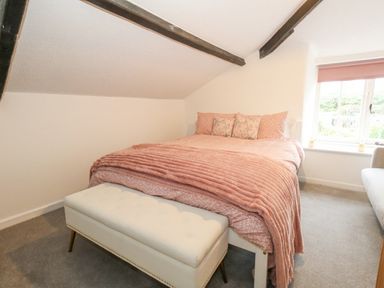 A bedroom with a bed covered with pink bedding a white bench at the foot of the bed and a window with a pink blind at Half Pint Cottage in Beaminster