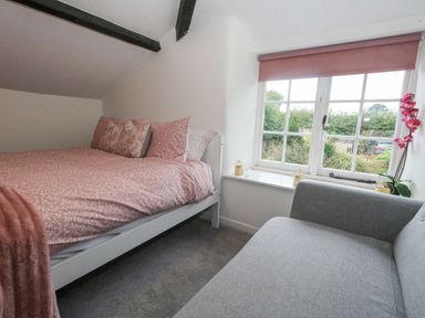 A bedroom with a bed with pink bedding and pillows next to a window with pink blind and a gray couch at Half Pint Cottage in Beaminster