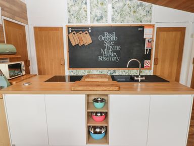 A kitchen island with cutting boards and mixing bowls in shelves a stove and sink behind it and a blackboard with herb names on the wall at The Bird House in Cenarth