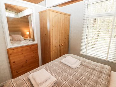 A bedroom with a wooden wardrobe a mirror two folded towels on a bed with a plaid blanket and a window with blinds at The Bird House in Cenarth