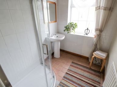 A bathroom with a shower enclosure a pedestal sink a window with a curtain a small stool with folded towels and a striped rug on the floor at Bofin in Oughterard County Galway