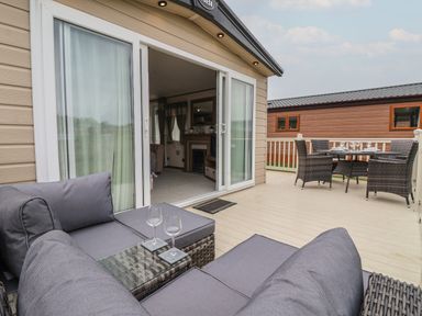 An outdoor deck with gray cushioned seating and a glass table with wine glasses adjacent to a dining table with chairs at Whispering Willows in Cayton Bay