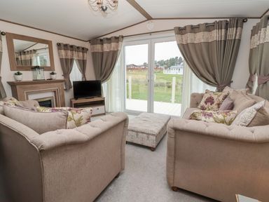 A living room with two beige sofas a patterned ottoman a TV on a wooden stand a fireplace with a mirror and windows with curtains at Whispering Willows in Cayton Bay