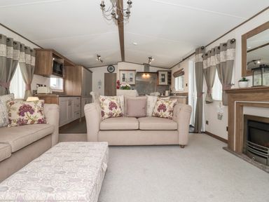 A living room with beige sofas and a patterned ottoman next to a kitchen and fireplace at Whispering Willows in Cayton Bay