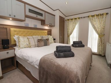 A bedroom with a large bed featuring multiple pillows and folded towels on top with floral curtains and overhead storage cabinets at Whispering Willows in Cayton Bay