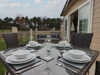 An outdoor dining table set with plates bowls glasses and cutlery with patio chairs and a sofa on a deck at Whispering Willows in Cayton Bay