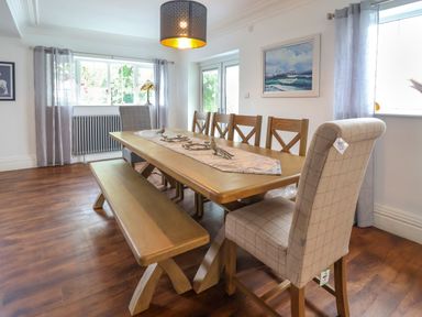 A dining room with a wooden table surrounded by chairs and a bench with windows and curtains at The Manor House Skerry Vore in Bryn Pydew near Llandudno Junction
