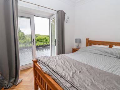 A bedroom with a wooden bed covered with striped bedding and gray curtains by glass doors opening to a balcony with trees visible outside at The Manor House Skerry Vore Bryn Pydew near Llandudno Junction