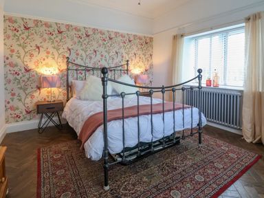 A bedroom with a metal bed frame a patterned rug wallpaper with birds nightstands with lamps and a window with curtains at The Manor House Skerry Vore Bryn Pydew near Llandudno Junction