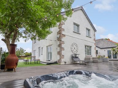 An outdoor area with a bubbling hot tub under a tree string lights bench table two lounge chairs a large clock on the wall and a white house at The Manor House Skerry Vore Bryn Pydew near Llandudno Junction