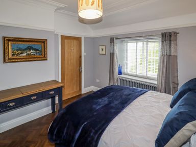 A bedroom with a bed covered in white and dark blue blankets a wooden door a blue console table a window with blinds and gray curtains and a framed painting on the wall at The Manor House Skerry Vore Bryn Pydew near Llandudno Junction