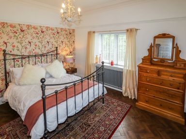 A bedroom with a metal bed frame a wooden dresser with a mirror a window with curtains and a decorative rug on the floor at The Manor House Skerry Vore Bryn Pydew near Llandudno Junction