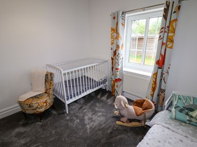A nursery room with a white crib a patterned chair with a throw and a plush rocking toy near a window with colorful curtains at The Manor House Skerry Vore Bryn Pydew near Llandudno Junction