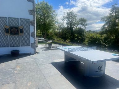 An outdoor patio with a ping pong table, dartboard on a wall, lounge chairs, and trees surrounding the area at The Manor House Skerry Vore in Bryn Pydew near Llandudno Junction