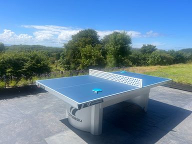 An outdoor blue table tennis table with two paddles on a stone patio with trees and grass in the background at The Manor House Skerry Vore Bryn Pydew near Llandudno Junction