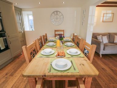 A dining room with a wooden table set with white plates bowls and cutlery eight chairs and a clock on the wall at Tillers Cottage in Ebberston