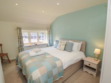 A bedroom with a double bed with pillows and folded towels a bedside table with a lamp curtains on windows and a wooden chair at Tillers Cottage in Ebberston