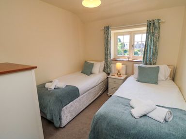 A bedroom with two single beds with blue throws and pillows a window with curtains and a nightstand with a lamp at Tillers Cottage in Ebberston