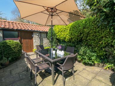 An outdoor patio with a table and six chairs under a large umbrella surrounded by greenery at Tillers Cottage in Ebberston