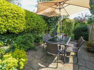 An outdoor patio area with a glass table and six wicker chairs under a large beige umbrella surrounded by green shrubs and plants at Tillers Cottage Ebberston