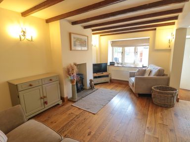 A living room with exposed ceiling beams a wood-burning stove a TV on a stand a sideboard two armchairs and a wicker basket at Tillers Cottage in Ebberston