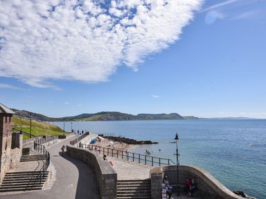 A seaside path with railings and steps leading to a small beach with people swimming and hills in the background at 16 Church Street in Lyme Regis