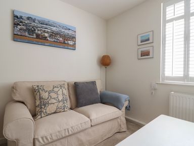 A beige sofa with cushions and a blue throw near a corner with a wicker floor lamp and framed pictures beside a window with white shutters at 16 Church Street in Lyme Regis