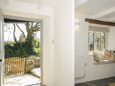 An open door leading to a garden with a wooden gate and a window seat with cushions inside a white room at Sunrise Cottage in St Kew Highway near Wadebridge